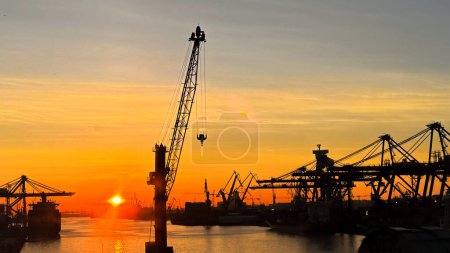 Sunset silhouettes cargo ships, cranes at sea port. Industrial loads freight at dock. Trade, transport infrastructure. Maritime logistics, global shipping. Clear sky reflects calm water