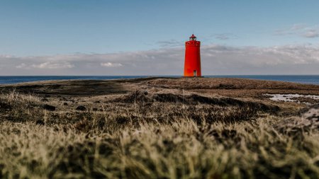 Un phare rouge surplombe une falaise rocheuse surplombant l'océan Atlantique, en Islande. Ciel bleu, avec quelques taches de nuages sombres visibles autour de la périphérie, ainsi qu'une dispersion.