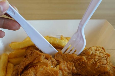 Téléchargez les photos : Fermer une cuillère à couverts en plastique en prenant un poulet frit croustillant avec des frites dans une boîte en papier sur une table en bois - en image libre de droit