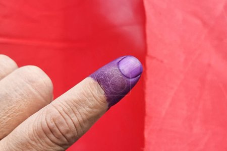 Téléchargez les photos : Gros plan d'encre violette indélébile sur l'index sur fond rouge. L'encre est utilisée lors des élections en Malaisie pour prévenir la fraude. - en image libre de droit