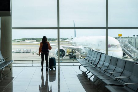 Foto de Mujer con la maleta en la mano, mira por la ventana del aeropuerto mientras espera su vuelo. - Imagen libre de derechos