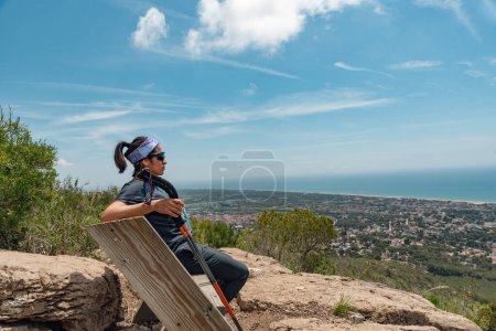 Woman takes a break on a mountaintop bench while gazing at the beauty of the Mediterranean coast.