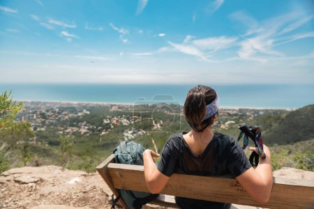 Hiker contemplating the Mediterranean coast, resting on a wooden bench at the top of the mountain.