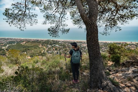 Hiker with a backpack and mountain sticks, contemplates the Mediterranean coast from above and below a tree.