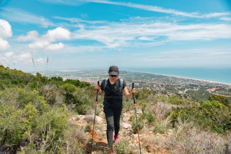 Woman supported by hiking poles continues on her way, while leaving behind the beautiful landscape of the Mediterranean coast.