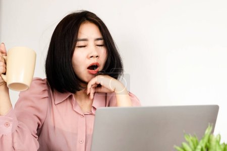 Téléchargez les photos : Fatigué asiatique femme bâillant et serrant une tasse de café alors qu'il est assis à son bureau - en image libre de droit