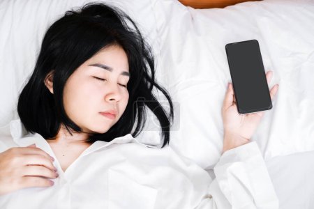 Foto de Concepto de la nomofobia con la mujer asiática durmiendo con la mano todavía embrague el teléfono móvil, simbolizando la constante necesidad de proximidad del teléfono - Imagen libre de derechos