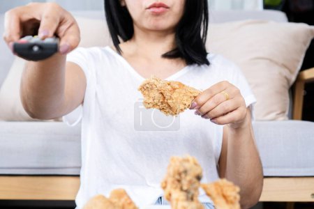 Photo for Lazy Asian woman eating fried chicken hand holding remote tv, unhealthy lifestyle concept - Royalty Free Image