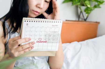 Téléchargez les photos : Souligné femme asiatique regardant calendrier ayant problème avec le cycle menstruel en attente pour le jour de l'ovulation - en image libre de droit
