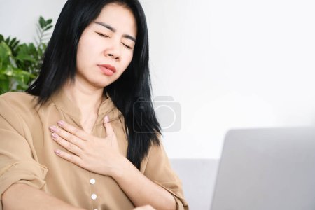 Téléchargez les photos : Femme asiatique souffrant de chagrin d'amour main tenant sa douleur thoracique tout en travaillant au travail assis au bureau essayer de respirer - en image libre de droit