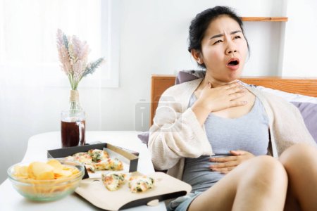 Foto de Mujer asiática que sufre de reflujo ácido después de comer comida chatarra en la cama se sienten incómodos y quieren vomitar - Imagen libre de derechos