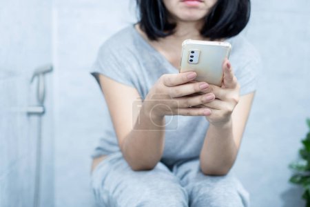Photo for Asian woman using mobile phone sitting too long in the toilet can cause constipation and hemorrhoids - Royalty Free Image