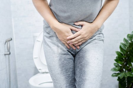 Stress Harninkontinenz, SUI mit einer Frau, die keinen Urin halten kann, Probleme mit der Blasenkontrolle hat, mit nassen Hosen im Badezimmer steht