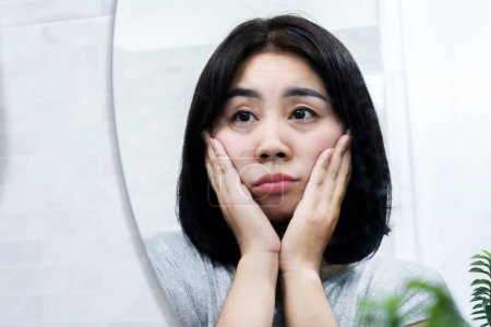 Téléchargez les photos : Femme asiatique regardant un miroir pour vérifier les problèmes de peau avec des sacs à yeux, des cernes, des rides et des pattes d'oie - en image libre de droit