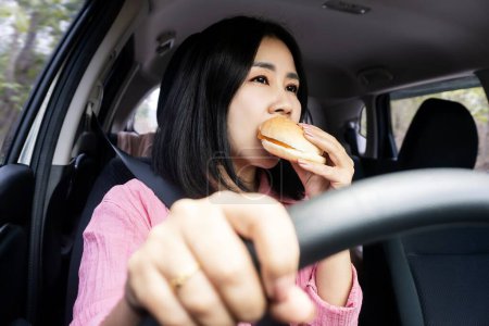Photo for Asian woman with bad eating habits is eating a burger while driving, which shows an unhealthy lifestyle because of junk food - Royalty Free Image