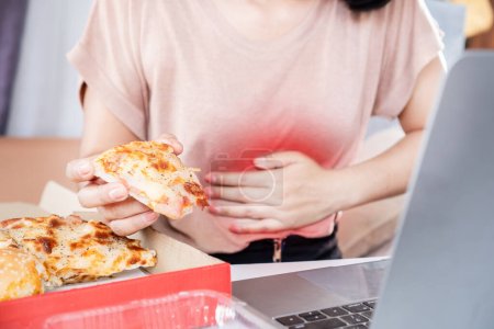 Frau leidet unter Magenschmerzen nach übermäßigem Verzehr von Junk-Food-Pizza am Schreibtisch Hand hält ihren Magen voller Gas, Keim von saurem Reflux