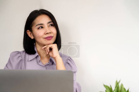 glücklich bei der Arbeit, asiatische Geschäftsfrau lächelt am Schreibtisch, schaut auf und denkt an Idee 