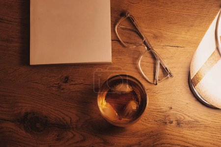 Téléchargez les photos : Bureau en chêne orné d'un verre de whisky, une ouverture, et la lueur ambiante d'une lampe de table - en image libre de droit
