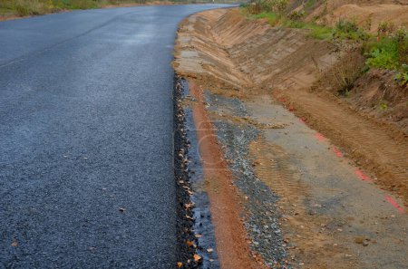 Photo for A newly built road, on the layer of which you can see successive layers of screed and asphalt coatings and penetration spraying. the edge is gradually shaped by earth at ditch, uncomplete, fall down - Royalty Free Image