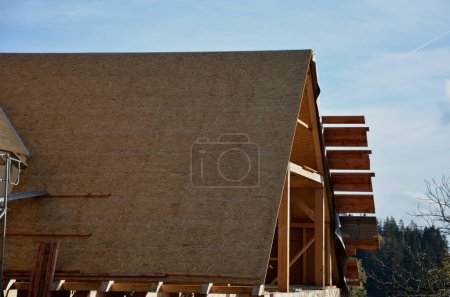 Photo for Wooden construction from assembled wooden beams, insulation with mineral wool and folding ceilings of walls with plywood and boards. roofers stand on the ridge and hammer nails, tape, insulated - Royalty Free Image
