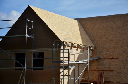 Photo for Wooden construction from assembled wooden beams, insulation with mineral wool and folding ceilings of walls with plywood and boards. roofers stand on the ridge and hammer nails, tape, insulated - Royalty Free Image