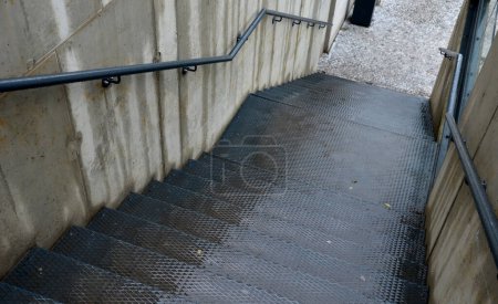 Foto de Paredes de hormigón entre las que se encuentra una escalera de metal con un patrón antideslizante. la barandilla sobre el hormigón expuesto está pintada de negro. - Imagen libre de derechos
