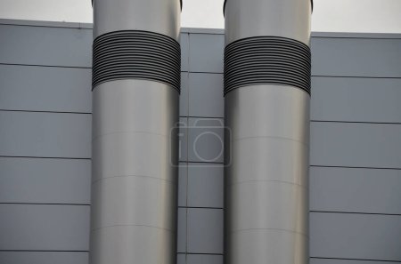Téléchargez les photos : Bâtiment industriel gris avec façade en tôle de lambris rectangulaires répétés. sur le mur sont deux cheminées argentées brillantes avec un tuyau en acier inoxydable courbé et biseauté. chauffage au gaz chaudière turbo - en image libre de droit