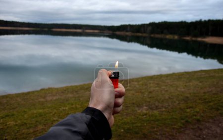 Téléchargez les photos : A man holds a lighter in his hand and burns gas in a small orange laminar by a lake with a forest. It gives a signal to the other side. fire calms and inspires pyromania. disposable red - en image libre de droit