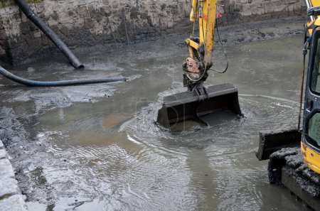 Foto de Removing sedimentary sludge from the bottom of a pond or a swimming lake in the village. the muddy water cleaning set with pumps vacuums the sump. a crawler excavator cleans bottom from deposits soil - Imagen libre de derechos