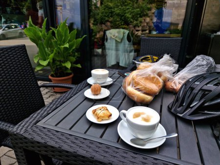 copieux petit déjeuner avec café et cappuccino à une table en plastique. le voyage à vélo commence par une collation dans la boulangerie. un casque de vélo gris repose sur la table