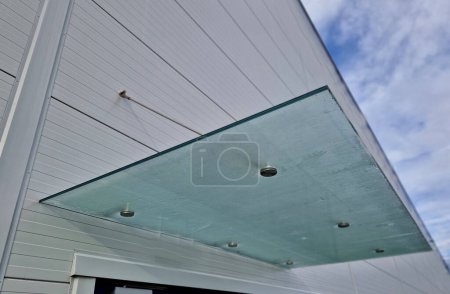 Glasdach über dem Eingang Industriegebäude mit Metallverkleidung der Fassade. dickwandige Glasscheibe hängt über der Tür an Metallstäben mit runden Scheibenhaltern, mattiert, Edelstahl, blau