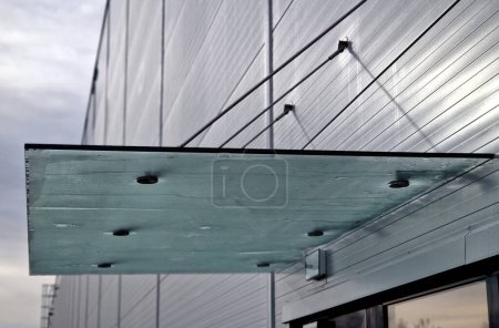 Glass roof above entrance industrial building with metal cladding of facade. thick-walled glass panel hangs above the door on metal rods with round targets holders, frosted, stainless steel, blue
