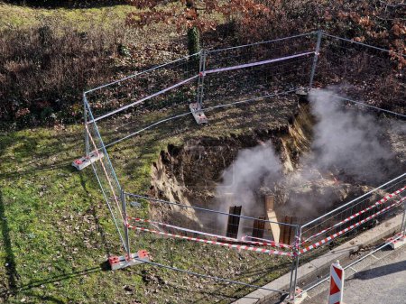 repairing pipes with hot water or steam in deep armored pit resembles a mine from which mist or dust is rolling. bounded by a temporary building fence as protection against the entry , tape barrier