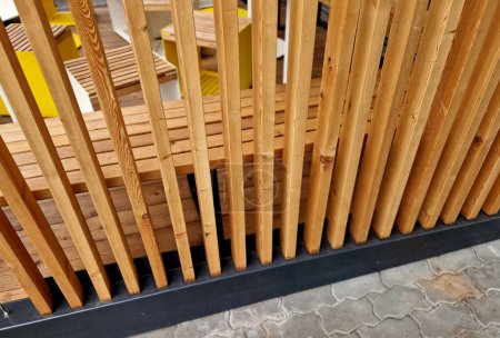 a pergola with slatted sides protects against the sun. a flowerpot with an evergreen buxus plant. seating in front of the cafe made of wooden elements, striped blinds and metal cables, bench