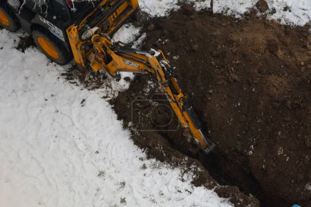 Téléchargez les photos : Tiraspol, Moldavie - 31 janvier 2023 : Une excavatrice JCB creuse une tranchée pour poser un tuyau d'égout par une journée nuageuse d'hiver. Mise au point sélective, faible profondeur de champ - en image libre de droit