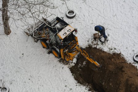 Photo for Tiraspol, Moldova - January 31, 2023: JCB excavator digs a trench for laying a sewer pipe. Worker of the water supply and sewerage service near the tractor on a cloudy winter day. Selective focus - Royalty Free Image