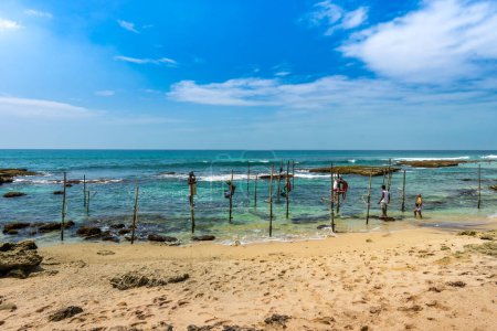Foto de Southwest Coast, Sri Lanka - 29 de enero de 2024: Los pescadores se equilibran hábilmente sobre pilotes de madera en medio de las aguas poco profundas, lanzando sus líneas en un método de pesca sereno y tradicional. - Imagen libre de derechos