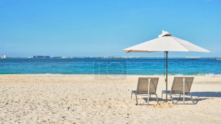 Foto de 2 sillas de playa y un paraguas blanco en las playas de Cancún en la arena blanca. en el fondo un océano azul turquesa y diferentes tonos de azul, pequeños edificios y un cielo lleno de nubes. - Imagen libre de derechos