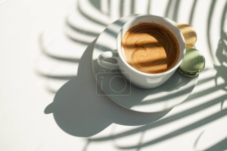 Coffee cup, top view. fresh black morning coffee with macaroni on a white wooden table and plant shadows.