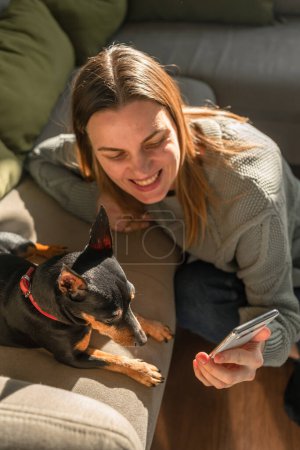 Photo for A woman shows a dog, a pinscher breed, a phone screen and smiles. A European woman and a pinscher on the sofa are looking at the phone screen - Royalty Free Image