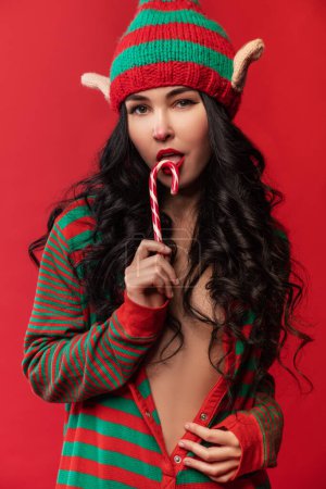 Photo for Sexy young woman in elf costume and with elven ears is standing on the red studio background and eating a striped candy - Royalty Free Image