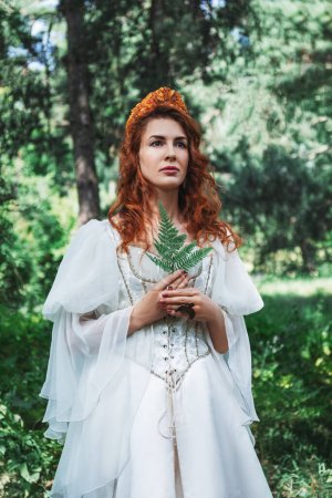 Téléchargez les photos : Belle femme aux cheveux roux dans un costume médiéval blanc, dans une couronne, avec un anneau d'aigle debout dans une forêt verte, princesse fantaisie - en image libre de droit