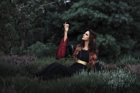 Foto de Hermosa princesa elfa oscura sentada en el césped con flores de brezo. Bruja de fantasía. - Imagen libre de derechos
