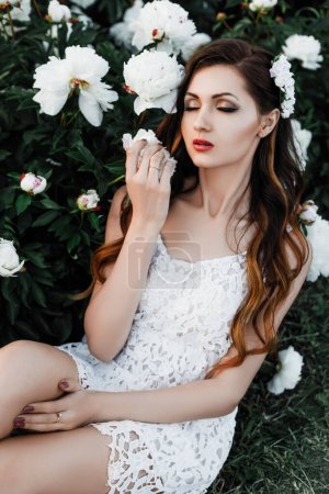 Téléchargez les photos : Portrait extérieur printemps d'une belle femme brune sexy aux cheveux bouclés, maquillage du soir, en robe blanche et avec des fleurs dans les cheveux, jardin, fleur, pivoines - en image libre de droit