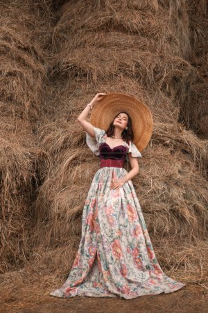 Photo for Beautiful young brunette woman in a long retro dress and a big straw hat standing near the hayloft - Royalty Free Image