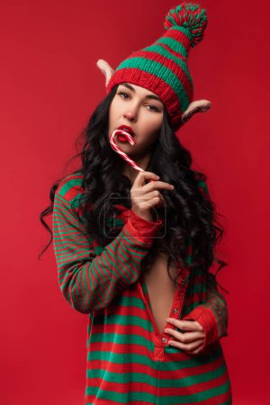Téléchargez les photos : Sexy jeune femme en costume d'elfe et avec des oreilles elfiques est debout sur le fond rouge studio et tenant un bonbon rayé - en image libre de droit
