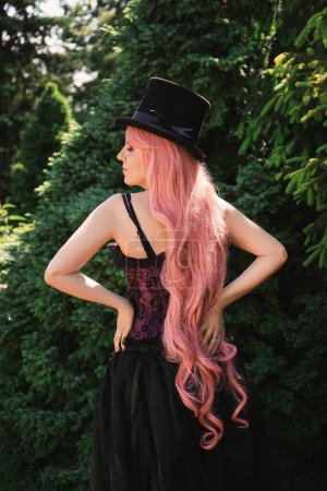 Foto de Hermosa mujer con el pelo rosa, en un sombrero de copa y en un largo vestido negro de noche con un corsé, se encuentra en el jardín en flores de azalea - Imagen libre de derechos