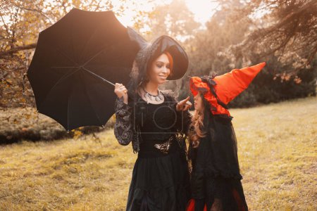 Foto de Niña bruja en vestido negro y rojo y sombrero puntiagudo con un paraguas en las manos, de pie delante de las viejas escaleras con hojas amarillas en traje de halloween - Imagen libre de derechos