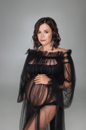Photo for Beautiful pregnant brunette woman in black transparent peignoir stands on gray studio background, looking at the camera - Royalty Free Image