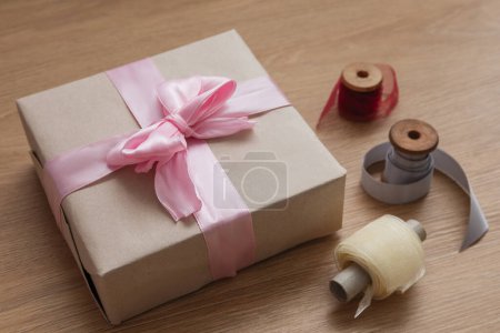 Foto de Regalo de cumpleaños en papel artesanal, con cinta rosa y tres carretes con cintas en la mesa de madera - Imagen libre de derechos